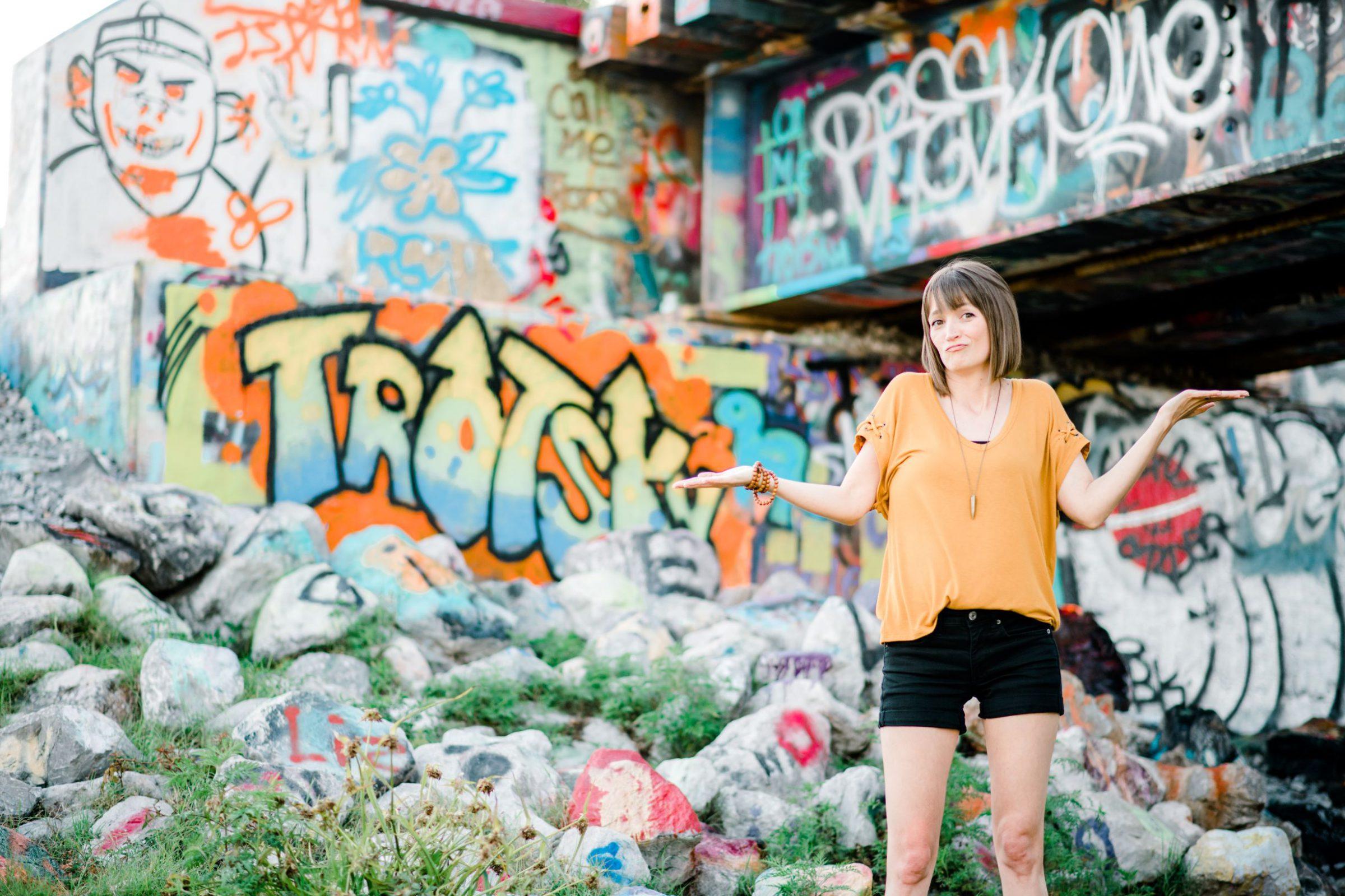 Tara standing outside with graffiti in the background wearing a yellow shirt and black shorts with her hands up looking unsure