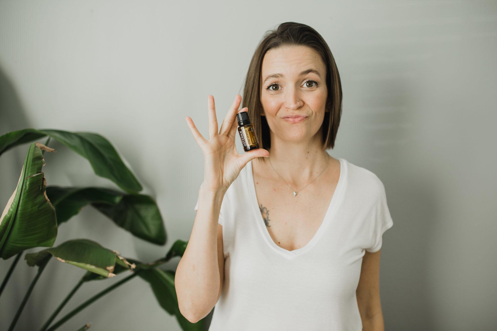 Tara wearing a white shirt, holding a Wild Orange essential oil bottle with white background and large house plant