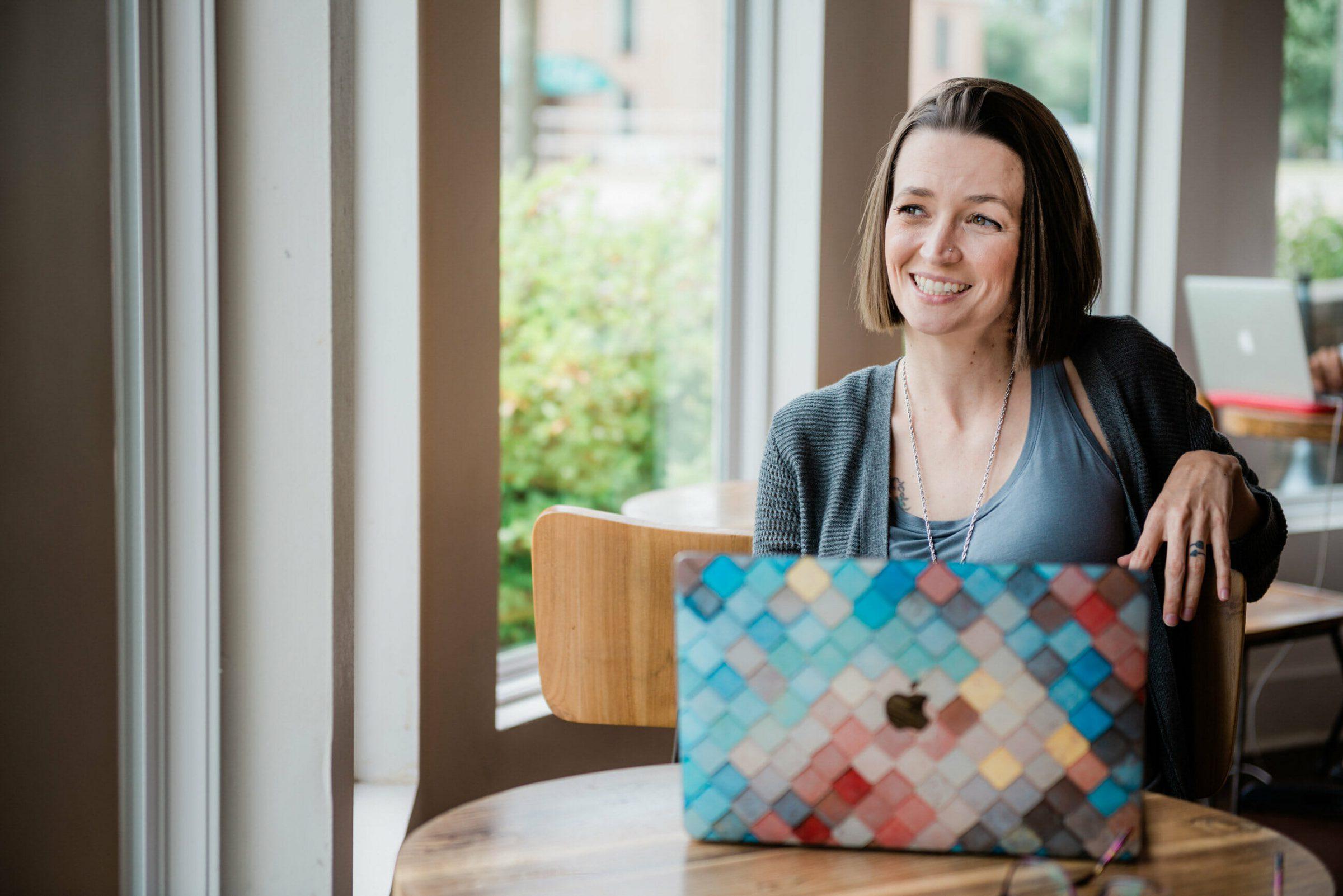 Tara sitting at window table in coffee shop, smiling on a laptop