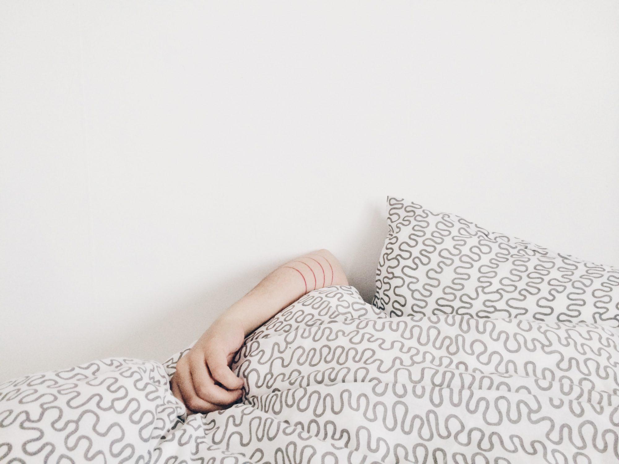 Woman laying in bed covering her face with white printed comforter