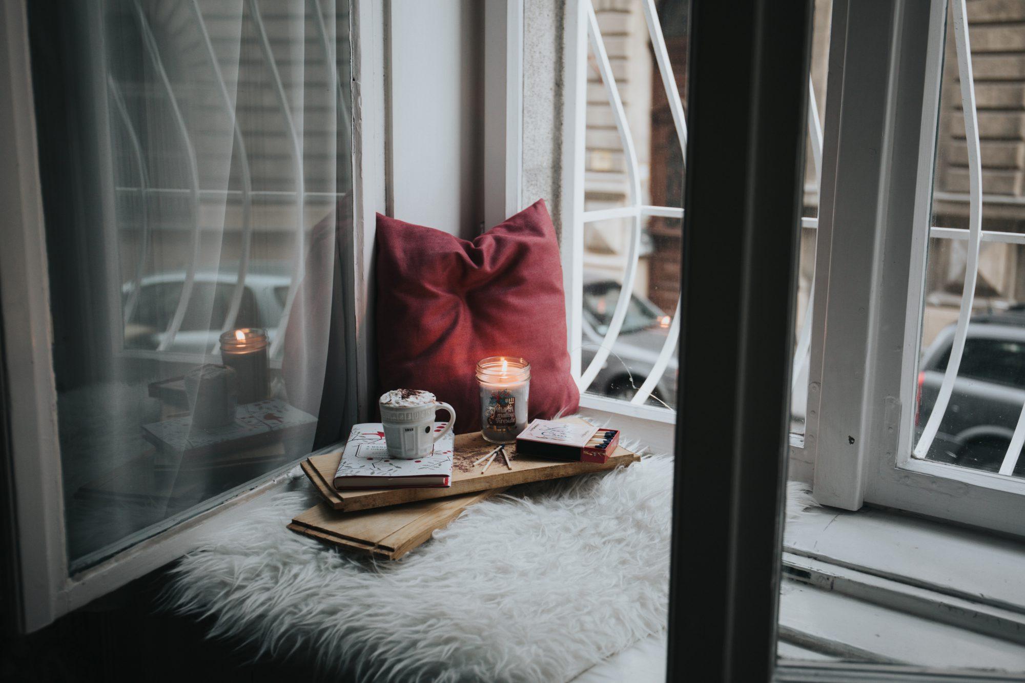 A comfy spot next to window sill with pillow, candle, a pad of paper and a mug.
