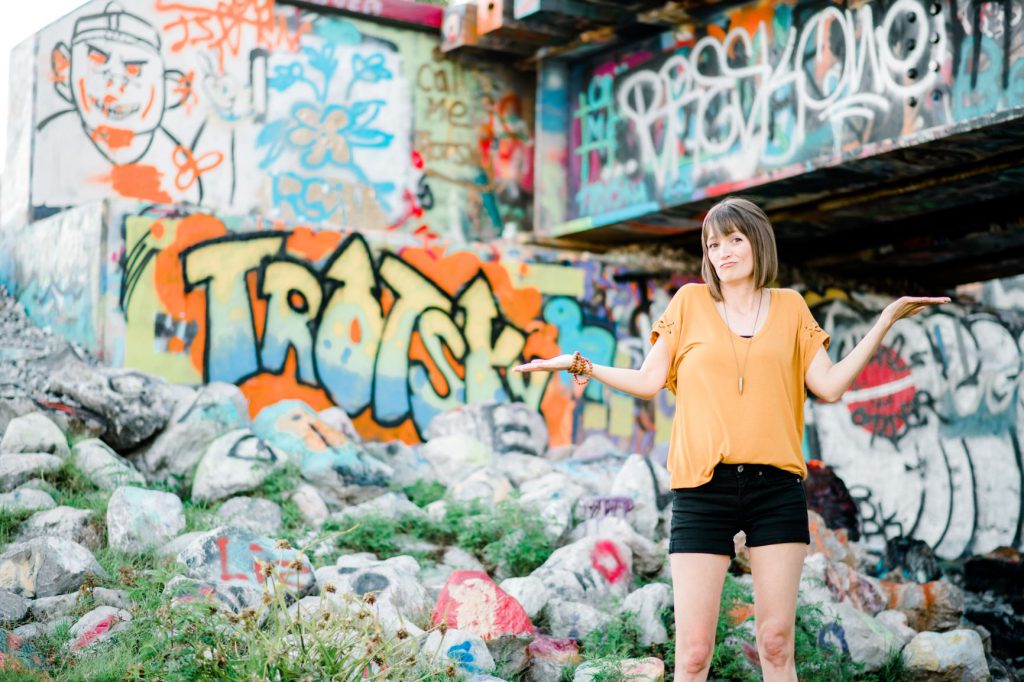 Tara in a yellow shirt and black shorts with arms out looking unsure, with a graffiti background.