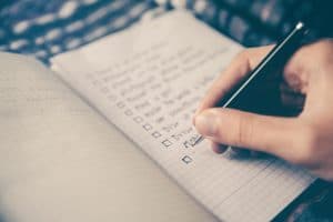 A notebook is open with a hand holding a pen writing a checklist.