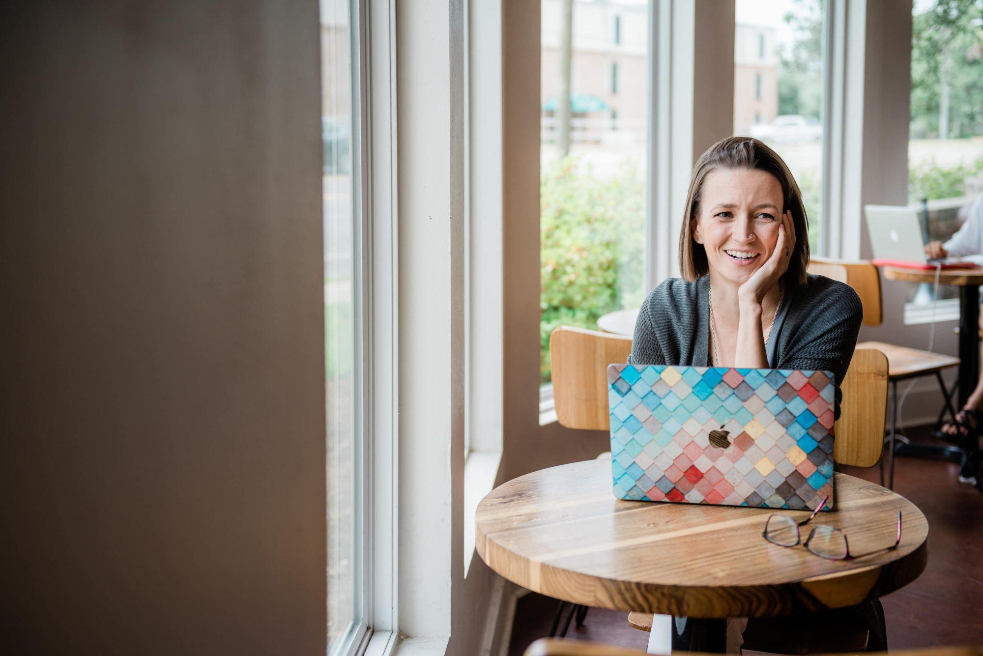 Tara sitting at wooden cafe table with laptop. Hand is placed on chin and face posed with a smile.