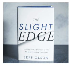 The book titled the Slight Edge by Jeff Olson. The title is in blue and black with a white hard copy cover. 