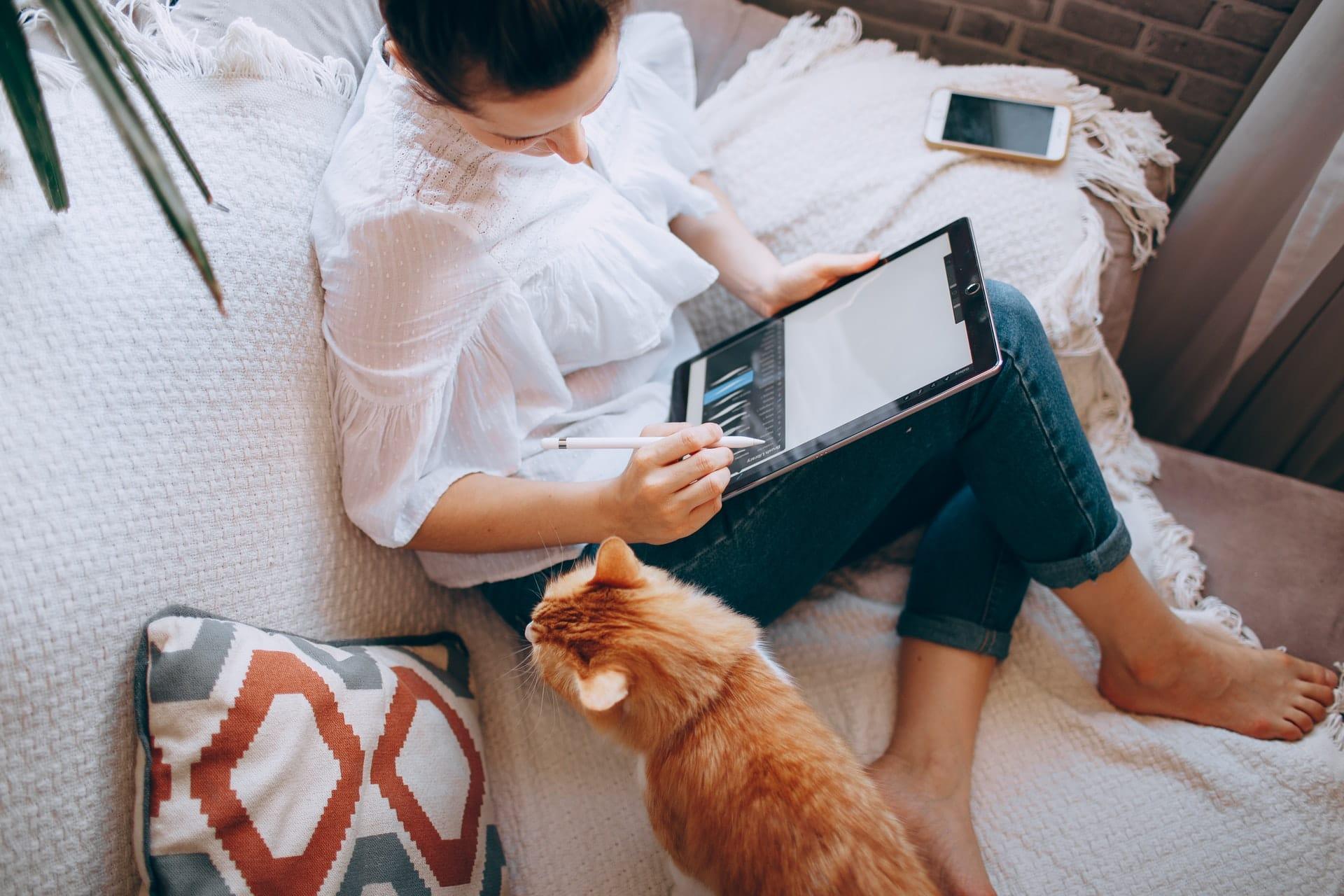 a young woman and her cat on the couch reading from her ipad