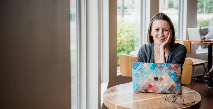 Tara sitting at wooden cafe table with laptop. Hand is placed on chin and face posed with a smile.