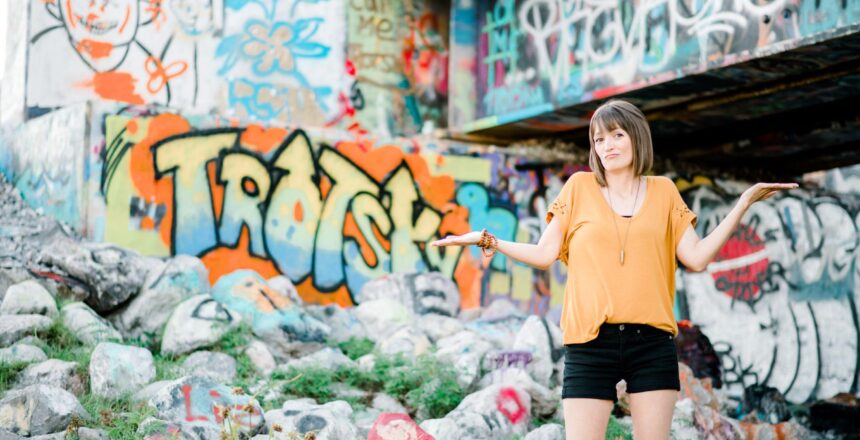 Tara standing outside with graffiti in the background wearing a yellow shirt and black shorts with her hands up looking unsure