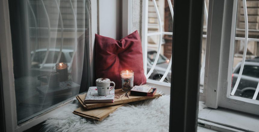 A comfy spot next to window sill with pillow, candle, a pad of paper and a mug.