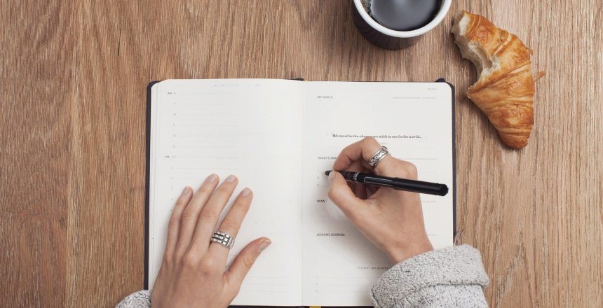 Open journal on wooden desk with female hands writing down business goals