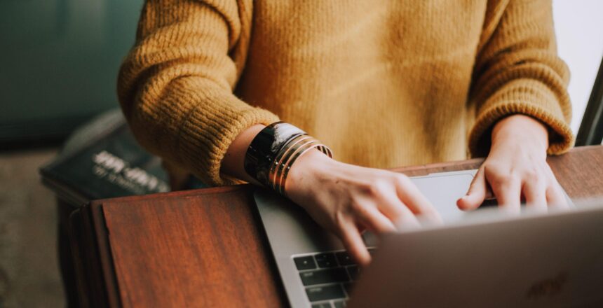 A woman with a gold bangle on right hand sitting at a wooden desk typing on a laptop.