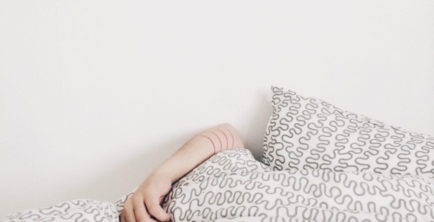 Woman laying in bed covering her face with white printed comforter