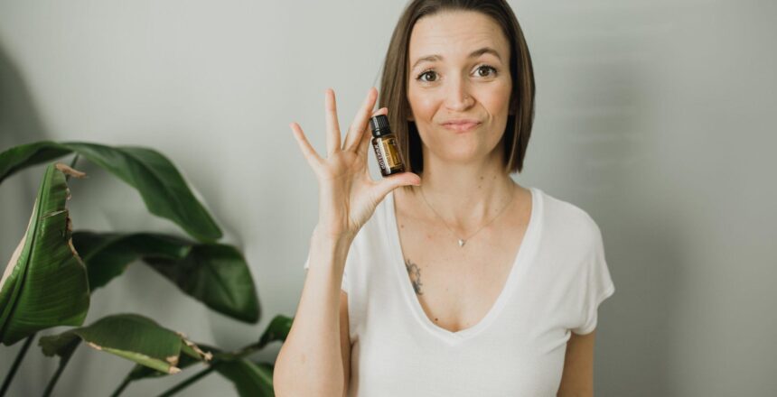 Tara wearing a white shirt, holding a Wild Orange essential oil bottle with white background and large house plant